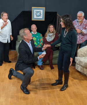 A group of people on stage watch as a man kneels to propose to a woman for the play On the Farce Day of Christmas for TRW Plays
