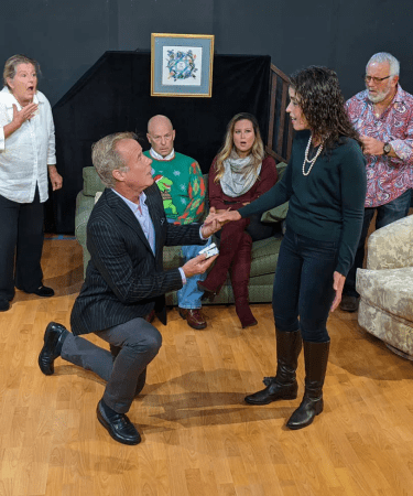 A group of people on stage watch as a man kneels to propose to a woman for the play On the Farce Day of Christmas for TRW Plays